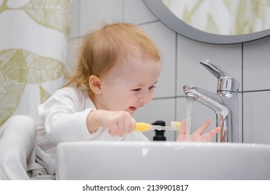 A Toddler Sitting On The Washbasin Cabinet And Brushing Teeth, Washing Tooth Brush In Water In Sink, Learning New Skills. Daily Morning Hygiene Routine. Personal And Teeth Hygiene.