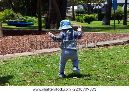 Toddler walking unsteady on feet in a park on a bright sunny day, wearing a hat, jumper and long pants.
