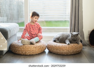 A Toddler Wearing Pink Pyjamas, Long Socks And Slippers Sits On A Wicker Stool And Looks At Her British Short Hair Cat Sat Next To A Her In A House In Edinburgh, Scotland, UK