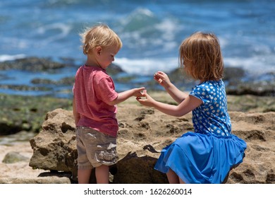 A toddler-aged boy and a preschool girl play together on a beach with water in the background, giving something back and forth