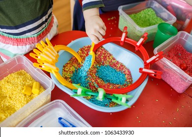 Toddlers playing with sensory bin with colourful rice on red table.