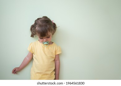 A Todler Girl Is Ashamed After A Childish Prank. A Child In A Yellow T-shirt Stands With A Pacifier Lowered Her Eyes Against The Background Of A Green Wall