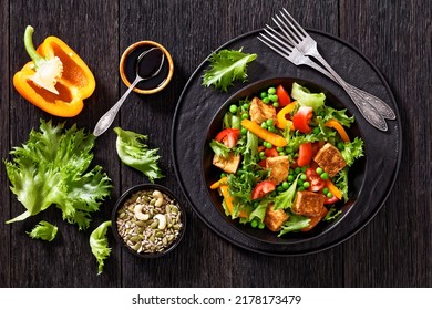 Tofu Salad With Crunchy Lettuce Leaves, Green Peas, Tomatoes, Yellow Sweet Pepper In Black Bowl With Dark Soy Sauce Dressing  And Mixed Seeds On Wooden Table, Horizontal View From Above, Flat Lay