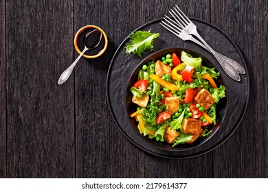 Tofu Salad With Crunchy Lettuce Leaves, Green Peas, Tomatoes, Yellow Sweet Pepper In Black Bowl With Dark Soy Sauce Dressing On Wooden Table, Horizontal View From Above, Flat Lay, Free Space