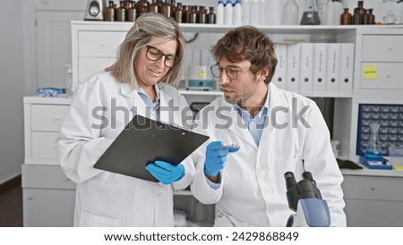 Together, two smiling scientists - a man and a woman, ardently scribble notes, sitting amidst bustling activity in the lab, medical samples in hand.