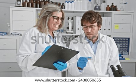 Together, two smiling scientists - a man and a woman, ardently scribble notes, sitting amidst bustling activity in the lab, medical samples in hand.