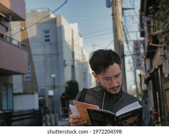 Tokyo, Japan - 02 04 2019: A Tourist In Japan Reads A Travel Guidebook. Young Tourist With Glasses Reads A Tokyo Guidebook To Orient Himself In The City. Old-fashioned Tourism Without A Smartphone.