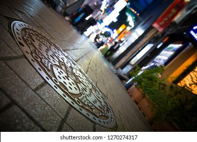 TOKYO, JAPAN - 23 JUNE 2016 - Decorative Manhole Cover And Low Street View In The Ikebukuro District Of Tokyo. Night Shot With The Neon Lights Of Local Cafes And Bars. INTENTIONAL 45 Degree Angle Shot