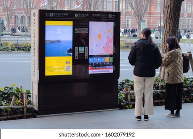 TOKYO, JAPAN - December 29, 2019: Visitors look at a touch screen tourist information board which includes a free Wifi service located on a sidewalk with the iconic Tokyo Station in the background.