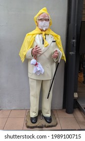 Tokyo, Japan - June 11, 2021: Colonel Sanders Statue Of A Kentucky Fried Chicken(KFC) Branch In Japan Is Wearing A Raincoat And A Facemask. He Is Also Holding A Small Handmade Doll Made Of White Cloth