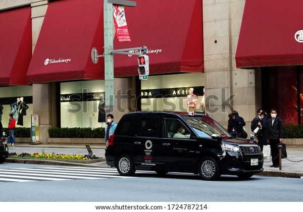 TOKYO, JAPAN - March 9, 2020: Taxi & people in\
front of Nihonbashi\'s Takashimaya store. The people wear face masks\
due to the coronavirus outbreak. The car is Toyota JPN Taxi with\
Tokyo Olympic logo.