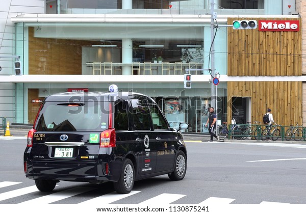 TOKYO, JAPAN - May 19, 2018: A taxi at junction in\
front of Aoyama\'s branch of German domestic appliance store Miele.\
The car is a Daiwa Taxi Toyota JPN Taxi with the Tokyo Olympic logo\
on it.
