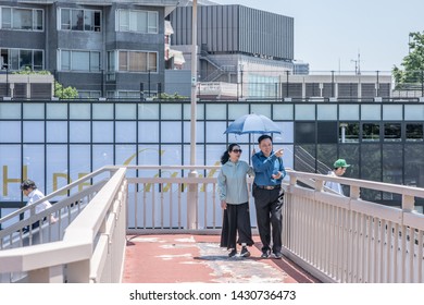 Tokyo, Japan - May 23, 2019: People Walk Across The Famous Harajuku Bridge In Shibuya, On A Sunny Day, Carrying An Umbrella For Sun Protection.