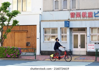 TOKYO, JAPAN - TOKYO, JAPAN - May 3, 2022: Cyclist on a bikeshare scene rented e-bicycle passing a music school in Tokyo's Koto Ward's Kiyosumi area. Some motion blur.