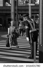 Tokyo, Japan - November 26, 2012: An Older Japanese Lady In Kimono / Traditional Dress Is Crossing At A Pedestrian Crossing While A Traffic Ward Is Bowing For Her. In Black And White