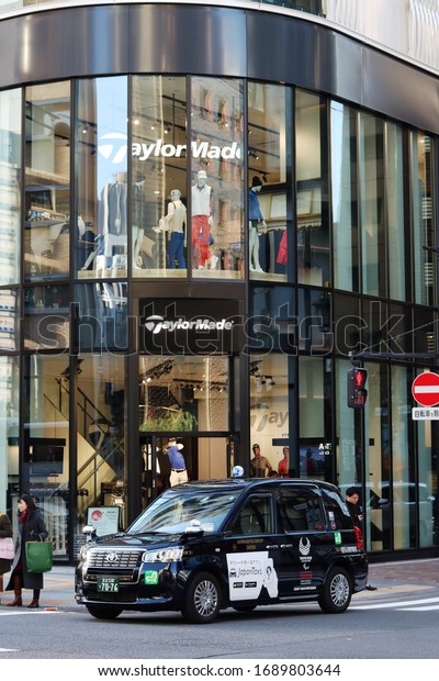 TOKYO, JAPAN - November 30,\
2019: A Toyota JPN Taxi with Tokyo Paralympic 2020 logo on it in a\
street in Ginza in front of a TaylorMade golf store. Some motion\
blur.
