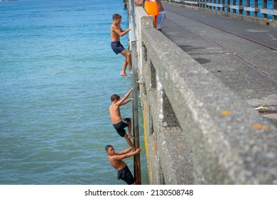 Tolaga Bay New Zealand - February 5 2022; Three Local Boys Climbing Up Ladder To Jump Of Tolaga Bay Wharf Under Encouragement Of Their Father.