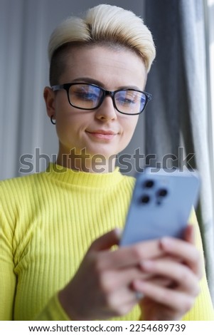 Tom boy female with short blond hair typing sms message on smartphone. Stylish white woman in glasses texting online with mobile phone
