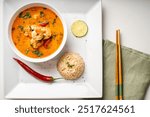 Tom Yam Kung Spicy Thai soup with shrimp and cilantro in a white bowl on a light background with rice and red hot pepper, lime on a square plate with chopsticks on a green napkin. High quality photo