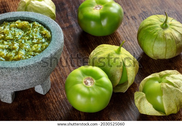 Tomatillos, green tomatoes, with salsa verde,\
green sauce, in a molcajete, traditional Mexican mortar, on a dark\
rustic wooden\
background