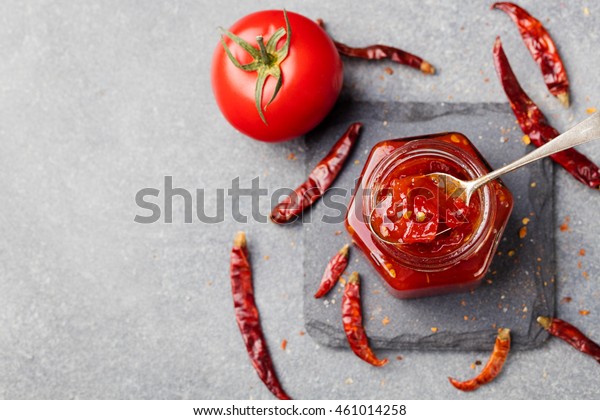 Tomato and chili sauce,\
jam, confiture in a glass jar on a grey stone background Copy space\
Top view