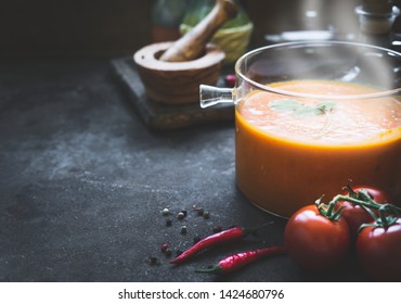 Tomato soup in glass cooking pot on dark kitchen table with ingredients. Copy space. Healthy food. Vegetarian eating
