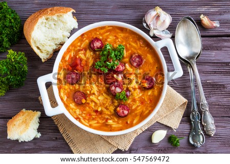 Tomato soup with orzo and smoked sausages in white casserole on wooden rustic table. Fresh bread and parsley, vintage spoon, top view.