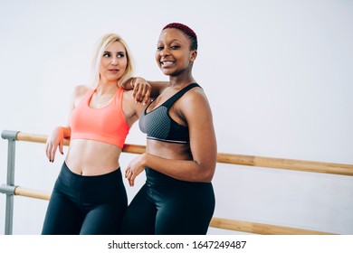 Toned sportswomen in activewear standing in fitness center room and leaning on ballet barre while blonde looking at smiling African American instructor