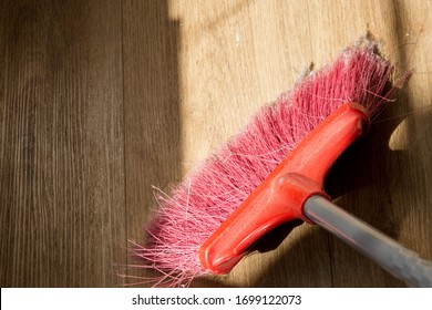 Tools For Cleaning The House. Red Sweeping Brush With Artificial Bristles And A Long Handle On The Background Of The Vinyl Floor Covering In The Kitchen, Imitated By Oak, Close-up