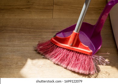 Tools For Cleaning The House. A Red Sweeping Brush With Artificial Bristles And A Purple Dustpan Against The Background Of The Vinyl Floor Covering In The Kitchen Close Up During Cleaning