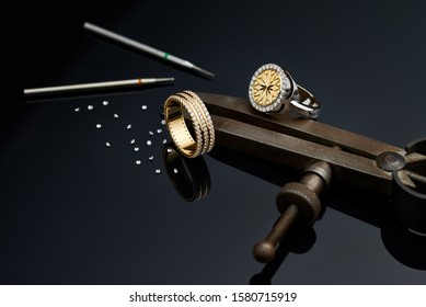 Tools and equipment for working on a black background. Two gold rings with diamonds and a scattering of precious stones. Engraver at work on diamond jewelry