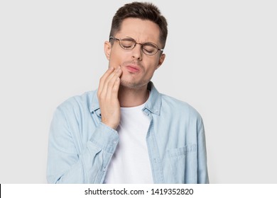 Tooth Ache Concept. Head Shot Studio Portrait On Grey Background Unhealthy Suffering Man In Glasses Jeans Shirt Feeling Teeth Pain, Guy Closed Eyes Holds His Cheek With Hand Painful Face Expressions