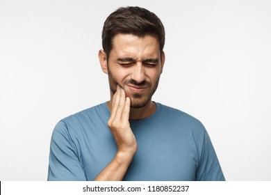 Tooth ache concept. Indoor shot of young male feeling pain, holding his cheek with hand, suffering from bad toothache, looking at camera with painful expression