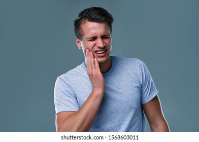 Tooth ache concept. Indoor shot of young male feeling pain, holding his cheek with hand, suffering from bad toothache, looking at camera with painful expression - Shutterstock ID 1568603011
