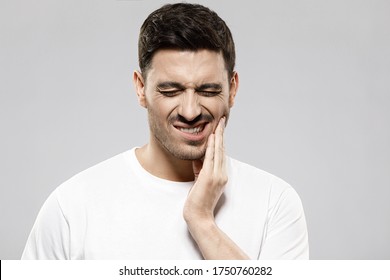 Toothache Concept. Portrait Of Young Man In White T-shirt Holding His Cheek Trying To Ease Severe Tooth Pain, Isolated On Gray Background