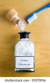 A Toothbrush And A  Bottle Of Sodium Bicarbonate (baking Soda) Laid Flat And Shot From Overhead On A Wooden Table.  Cork Open And Powder Spilling On To Table.