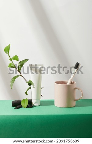 A toothbrush is placed into the pink cup. Green tea branch and bamboo charcoal are placed next to an empty cosmetic tube on a green table. White background with shadow. Branding and advertising.