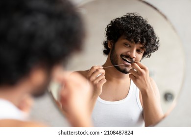 Toothcare Concept. Bearded Indian Man Using Teeth Floss Caring For Oral Hygiene And Dental Health, Looking At Reflection In Mirror In Bathroom. Guy Cleaning And Flossing Teeth.