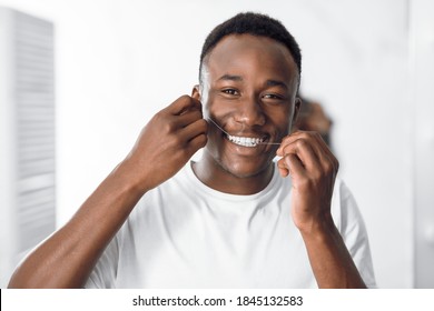 Toothcare And Hygiene. African American Guy Flossing Teeth Using Floss Smiling To Camera Standing In Modern Bathroom At Home. Healthy Teeth Cleaning Routine Concept