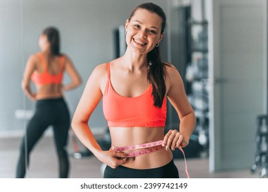 Toothy smiling American girl measuring waist happily smiling at sport club standing against blurry gym, shelve with fitness equipment. Healthy lifestyle. - Powered by Shutterstock