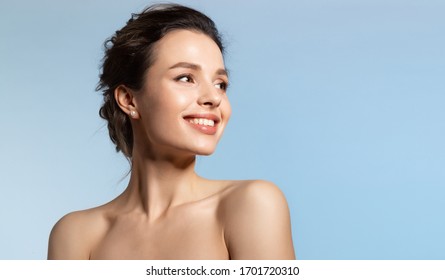 Toothy Smiling Young Woman With Shiny Glowing Perfect Facial Skin And Bare Shoulder Looking Aside Beauty Studio Portrait On Blue Copy Space. Cosmetology, Dermatology. Dental And Skin Treatment