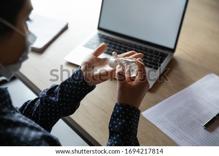 Top above shoulder view young cautious indian girl in protective facemask using antibacterial gel on hands before starting work on computer in office, stop prevent spreading corona virus infection.