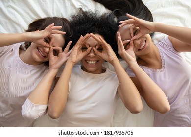 Top above view three happy multiracial diverse young pretty sincere women lying on bed in pajamas, making funny grimaces binoculars with fingers, having fun together at sleepover hen girls party. - Powered by Shutterstock