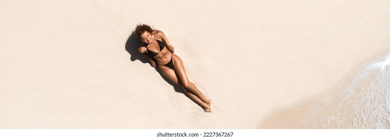 Top aerial drone view of woman in swimsuit relaxing and sunbathing on beach white sand near the ocean. Attractive brunette girl in black bikini laying on a sandy coast with palm leaf. Gorgeous tanned
