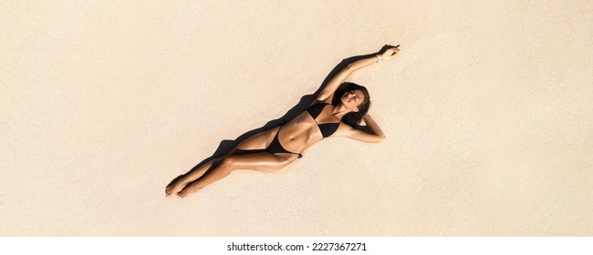 Top aerial drone view of woman in swimsuit relaxing and sunbathing on beach white sand near the ocean. Attractive brunette girl in black bikini laying on a sandy coast with palm leaf. Gorgeous tanned