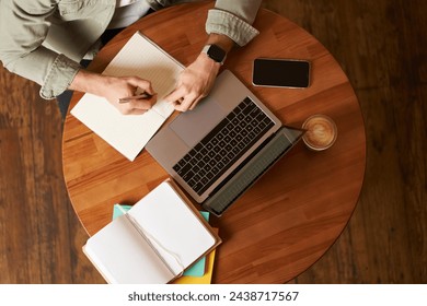 Top aerial view of male hands taking notes, writing in notebook, man sitting at round desk in cafe, working on laptop, studying. - Powered by Shutterstock