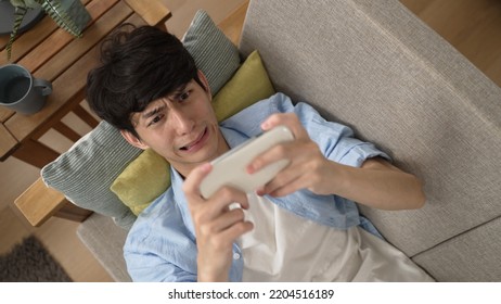 Top Angle Shot Of A Frustrated Asian Guy Lying On Sofa And Looking At His Mobile Phone With A Sad Face After Losing The Exciting Game In The Living Room At Home