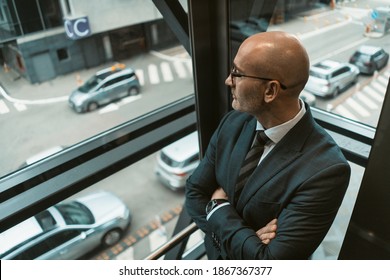 Top Angle View. Contemplative Bald Businessman In Suit And Glasses Looking Away In The Window Or Glass Elevator With A Busy Street View. Pensive Business Man Looking Outside Of The Window. 