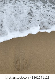 Top angle view of soft ocean wave foam washing over footprints on a sandy beach.