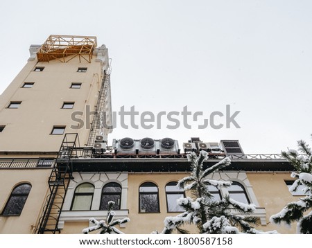 Top of the building with a tower, windows and fire ladders with snow-covered fir trees in the foreground. Bottom up view